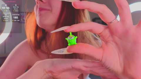 A video shows a young woman with long red hair and fair skin, holding a small, green, cartoonish figure. She has long, manicured nails and a silver ring on her right hand. The background is blurred with a circular display showing 10:50, 170, and a graph.