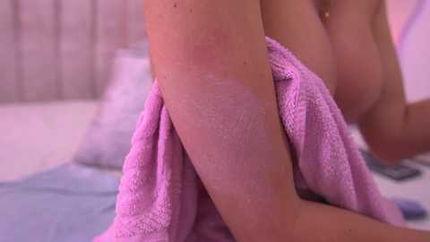 Video of a fair-skinned woman's upper thigh and leg, covered with a soft, pink towel, on a bed with blue sheets and pillows, in a softly lit room.