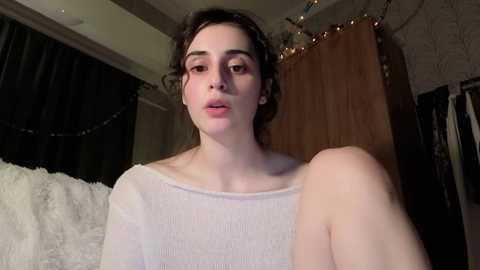 A video of a fair-skinned woman with curly dark hair, wearing a white knit sweater, sitting indoors, with a cozy, dimly lit room in the background.