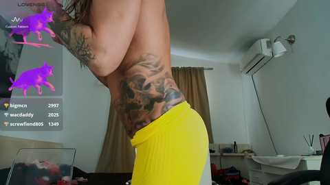 A video of a topless woman with large breasts and extensive tattoos on her side, wearing yellow shorts, standing in a modern living room with beige walls and an air conditioner.