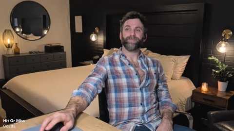 Video of a bearded, middle-aged man with short brown hair in a plaid shirt, seated in a modern bedroom with dark wood furniture, beige bedding, and two bedside lamps.