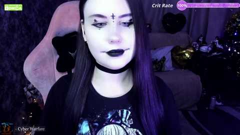A video of a pale-skinned woman with long black hair and dark makeup, wearing a black choker and a graphic T-shirt, seated in a dark room with a gaming setup.