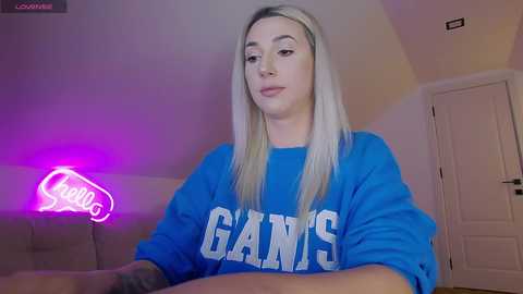 Video of a young, slim, blonde woman with fair skin wearing a blue \"GAINS\" sweatshirt in a dimly lit room with a pink neon sign and white door.