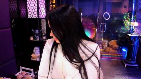 Video of a woman with long black hair in a white coat, standing near a black shelf with a teddy bear, colorful Christmas lights, and a window displaying a festive cityscape at night.