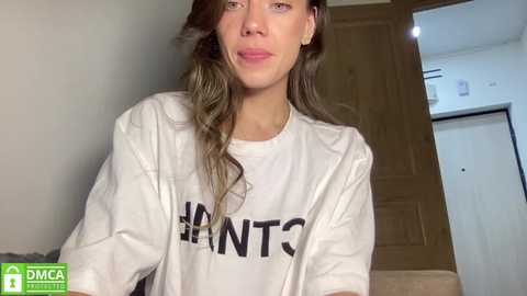 Video of a young woman with long, wavy brown hair, wearing a white t-shirt with \"HUNTS\" printed in black, standing indoors near a wooden door and a white wall.