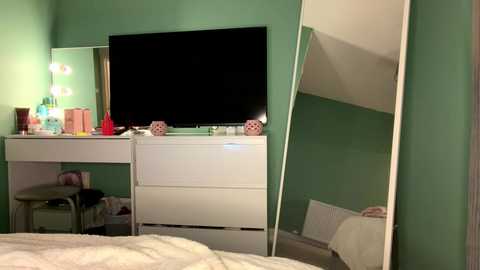 Video of a cozy bedroom with green walls, featuring a white dresser, TV, mirror, makeup table, and a bed with white bedding.