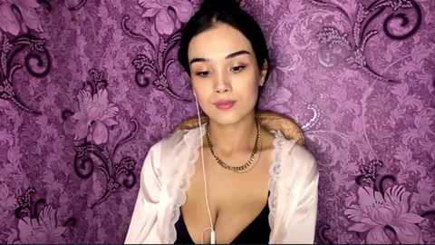 Video of a young woman with medium skin tone, dark hair pulled back, wearing a sheer white blouse, black bra, gold chain necklace, and a purple floral-patterned background.