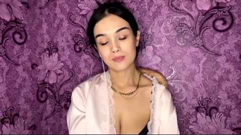 Video of a young woman with light skin, dark hair, and a slender physique, wearing a light pink satin robe, against a purple floral-patterned background.