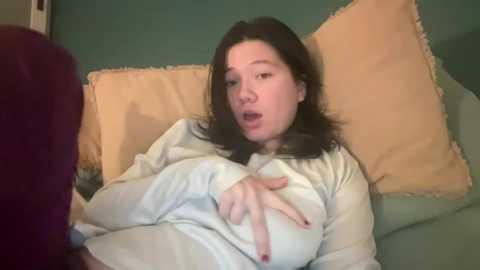 A video of a fair-skinned woman with shoulder-length dark hair, wearing a light sweater, making a peace sign on a couch with teal and yellow pillows.