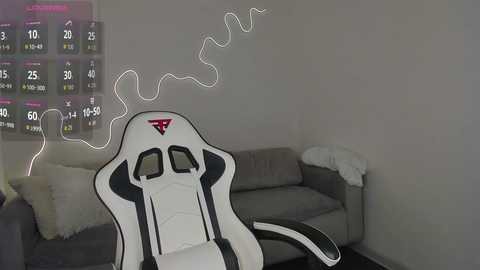 Video of a white gaming chair with a black and red logo, positioned in a minimalist living room with a gray sofa, surrounded by a digital gaming interface displayed on the wall.