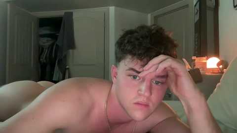 Video of a shirtless young man with curly brown hair, lying on his stomach in a dimly lit bedroom, looking tired, with a lamp and open closet door in the background.