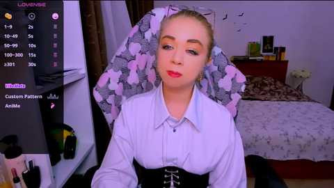 Video of a blonde woman with pale skin, wearing a white blouse and black corset, seated in a floral headwrap. Bedroom background with a bed, shelves, and a TV screen.