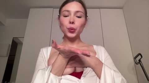 Video of a fair-skinned, young woman with brown hair, wearing a white robe, blowing kisses. Background features white cabinets and a door, creating a simple, modern interior setting.