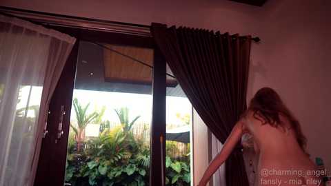 A video of a woman with light skin and brown hair, topless, adjusting dark curtains in a bedroom with a green garden view outside.