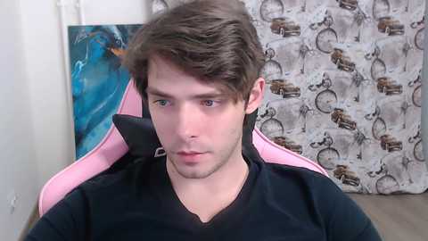Video of a young man with short brown hair and light skin, wearing a black V-neck shirt, sitting in a pink gaming chair against a wall with a patterned fabric.