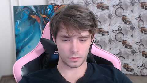 Video of a young white man with short brown hair, wearing a black shirt, seated in a pink gaming chair, with abstract and patterned wallpaper in the background.