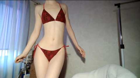 Media: Video of a slender, fair-skinned woman wearing a red, lacy bikini with side ties, standing in a dimly lit room with a bed, a wicker basket, and a standing lamp in the background.