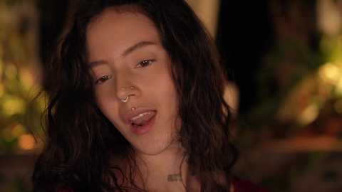 A video of a young woman with medium-brown skin, long dark hair, and a nose ring, smiling seductively in a dimly lit forest setting.