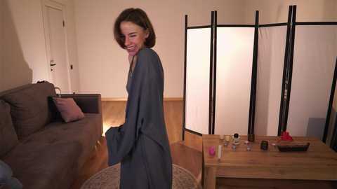 Media: Video of a woman in a gray robe laughing, standing in a warmly lit living room with a brown sofa, wooden floor, and decorative items.
