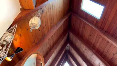 A video of a wooden interior with a triangular ceiling, featuring a round mirror, woven basket, and a skylight. The ceiling is richly stained with natural wood tones, creating a warm, rustic ambiance.