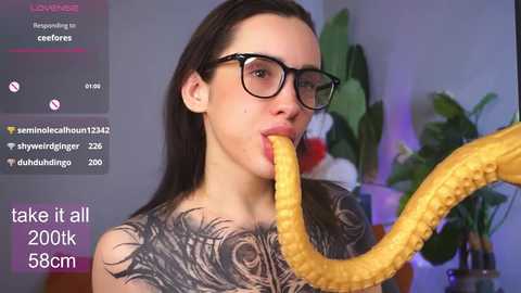 Video of a tattooed woman with glasses, eating a banana, in a room with a green plant.