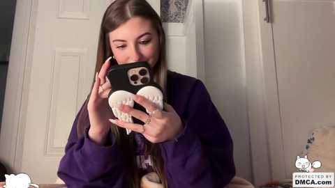 Video of a young woman with long brown hair, wearing a purple hoodie, taking a selfie in a bathroom, using a black phone with a white shark case.