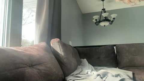 Media: Video of a cozy, lived-in corner of a room featuring a dark brown, plush leather sofa with a white blanket draped over it. A chandelier with white lightbulbs hangs from a ceiling with a pale greenish-gray paint.