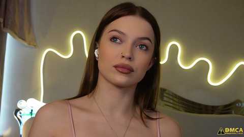 Video of a young woman with fair skin, blue eyes, and long brown hair, wearing a light pink spaghetti-strap top, with a glowing neon \"wave\" sign in the background.