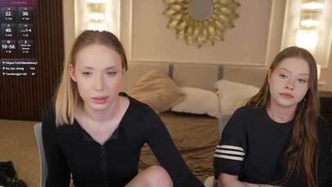 Video of two young women with fair skin and long, straight hair, one blonde and one brunette, wearing black tops, seated indoors. Background includes a bed with beige bedding, a wall mirror, and a digital clock.