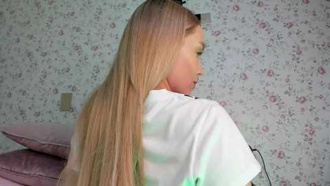 Video of a blonde-haired young girl with long, straight hair, wearing a white shirt, sitting on a pink bed, with floral wallpaper in the background.
