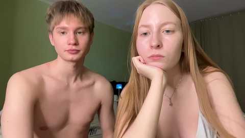 Video of two young, light-skinned individuals with fair skin and long hair. The man is shirtless, looking bored, while the woman, wearing a sleeveless top, rests her chin on her hand, appearing contemplative. They sit in a room with green walls and a TV in the background.