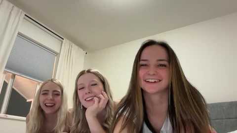 Video of three young women with long hair, smiling joyfully in a bright room with white walls and windows.