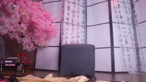 Video of a serene room with a dark gray ottoman, cherry blossom tree in a vase, and a shoji screen adorned with Japanese characters.