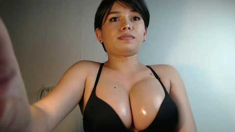 A close-up video of a young, short-haired woman with fair skin and medium-sized breasts in a black bra. She has a neutral expression and is indoors with plain walls in the background.