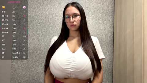 Video of a young woman with long black hair, wearing glasses and a tight white crop top, standing in front of a calendar.