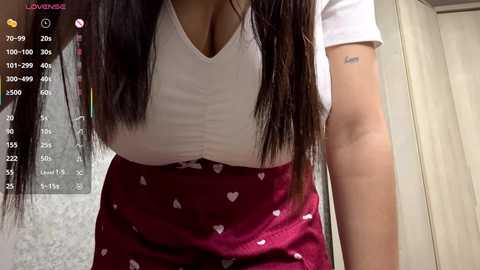 Video of a woman with long, straight dark hair, wearing a white V-neck shirt and maroon skirt with heart patterns, standing indoors.