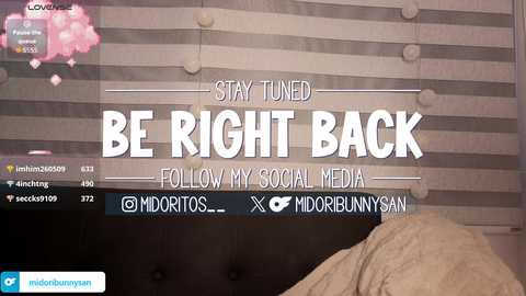 Video of a bedroom with a digital screen displaying \"Stay Tuned,\" \"Be Right Back,\" \"Follow My Social Media,\" and \"Midori.\" The background features a striped wall and a beige bed.