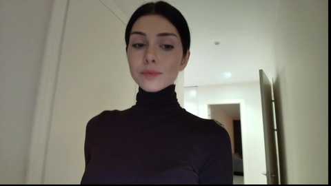 A video of a fair-skinned woman with dark hair, wearing a black turtleneck sweater, standing in a modern, dimly lit hallway with white walls and recessed lighting.