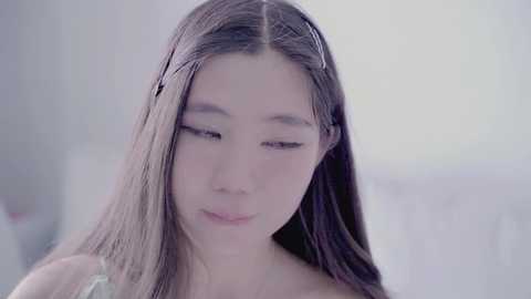 Media: A video of a young Asian woman with long, straight brown hair, wearing a light-colored top, softly smiling while gently touching her hair. The background is softly blurred in a neutral, pastel color palette.