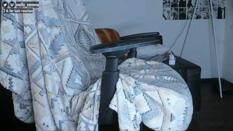 Video of a messy bedroom with a beige geometric-patterned blanket draped over a black office chair, white wall with black-and-white art collage, and cluttered floor.