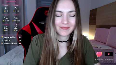 A video of a young woman with long, straight brown hair, smiling, in a green shirt, sitting in a red gaming chair, in a dimly-lit bedroom.