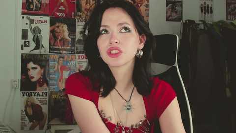 Video of a pale-skinned woman with dark hair, wearing a red dress, large earrings, and a necklace, sitting in a chair in a room adorned with magazine covers featuring Marilyn Monroe.
