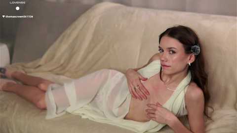 Video of a slim, fair-skinned woman with long brown hair, wearing a sheer, white, draped dress, lying on a beige couch. She covers her small breasts with her hands.