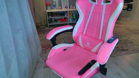 Media: Video of a pink and white gaming chair with adjustable armrests and headrest, placed on a wooden floor in a room with beige walls, a white shelf with toys, and a patterned rug.