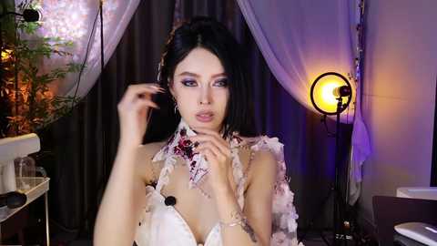 Video of a young East Asian woman with dark hair, wearing a white, floral-embellished dress, seated at a dimly lit table, against a dark backdrop with hanging white drapes.