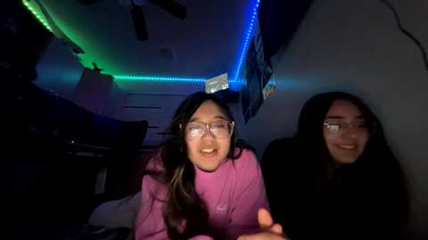 Video of two young women with glasses, one in a pink hoodie, the other in a black hoodie, sitting in a dimly lit room with green and blue LED strip lights, smiling and looking at something off-camera.