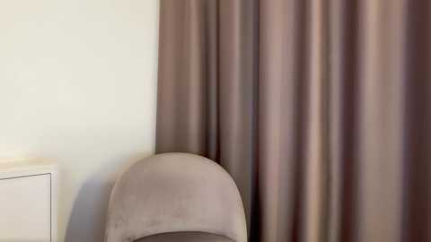 A video of a modern, beige upholstered chair with a soft, velvety texture, placed against a backdrop of soft, muted taupe drapes. The scene features a minimalistic, softly lit interior with a white nightstand visible in the left background.