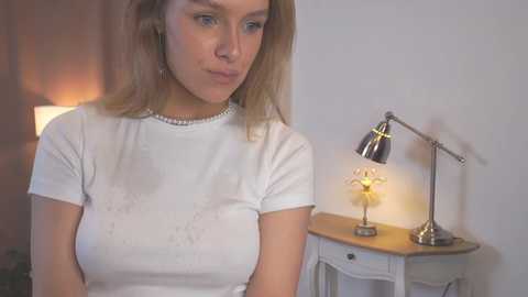 Video of a Caucasian woman with light brown hair, wearing a white T-shirt and pearl necklace, standing in a softly lit room with a table lamp and a small floral arrangement.