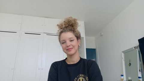 Video of a young woman with curly, blonde hair in a messy bun, wearing a black t-shirt with a logo, smiling in a minimalist, white-walled room with white cabinets and a mirror.