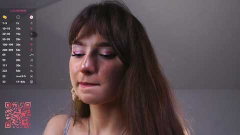 Video of a young woman with long brown hair, wearing pink eye makeup, standing indoors with a blurred background.